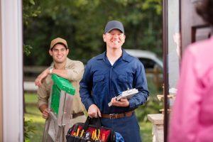 Caucasian and Hispanic repairmen or blue collar/service industry workers make service/house call at customer's front door. He holds his clipboard and tool box filled with work tools. Other worker holds ladder. Customer foreground.  Inspector, exterminator, electrician.  Service truck seen in background parked on road.